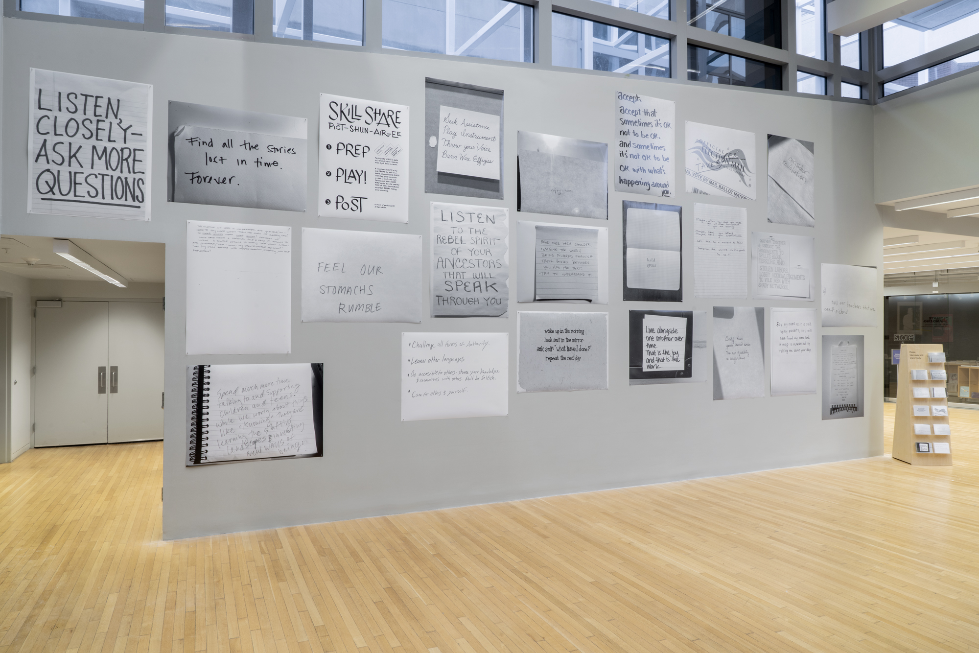 23 large format black and white posters of hand written text instructions by artists installed on an exhibition wall with a custom postcard stand next to it, displaying postcard versions of each poster, available to take away