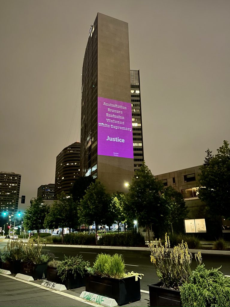 light projected text piece on tall facade of building in downtown Oakland, which in white on flourescent pink background reads in struck through text, "Assimilation / Erasure / Exclusion / Violence / White Supremacy" and on the last line in slightly larger text and no strikethrough, "Justice." At the bottom in smaller text reads Related Tactics.