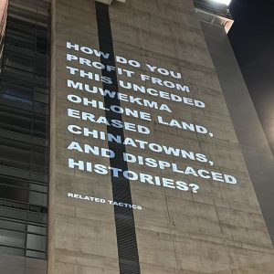 White text projected on a tall civic building reading, "How do you profit from this unceded Muwekma Ohlone land, erased Chinatowns, and displaced histories?"
